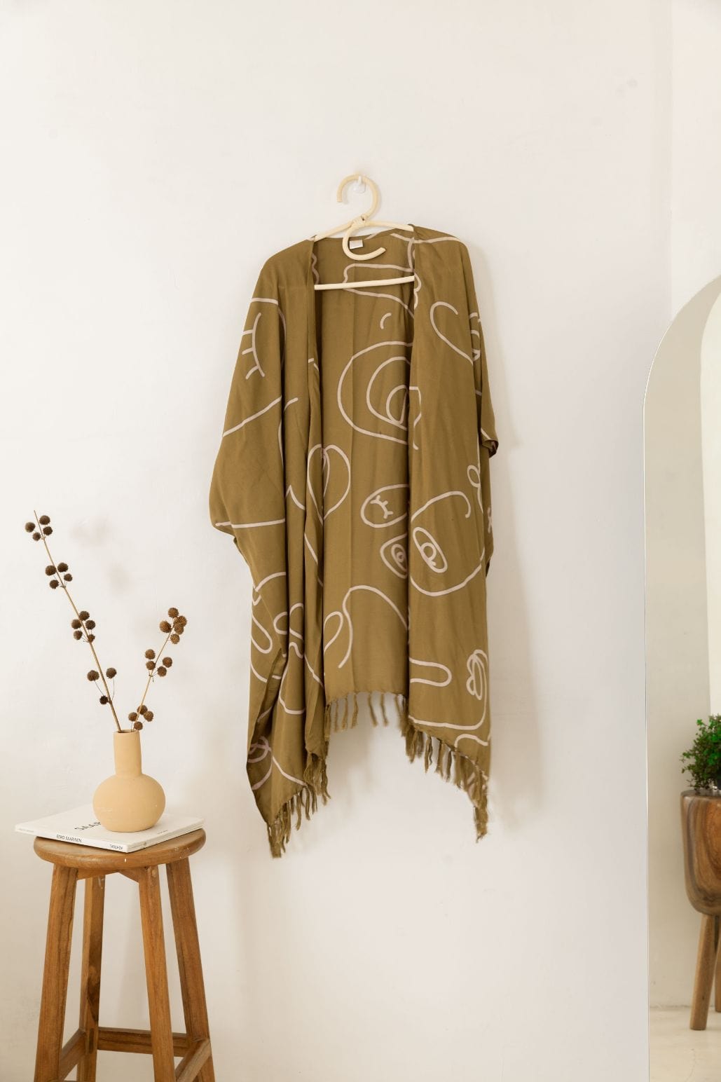 Village Thrive Faces Kimono, olive gold rayon, hanging on a wooden hanger against a wall.