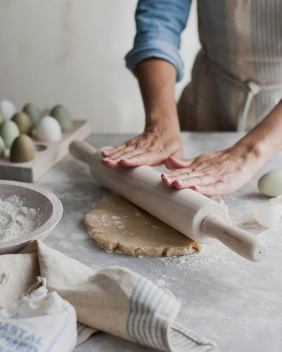 Baker pressing both hands on a whitewashed ash rolling pin while flattening dough on a flour-covered work surface.