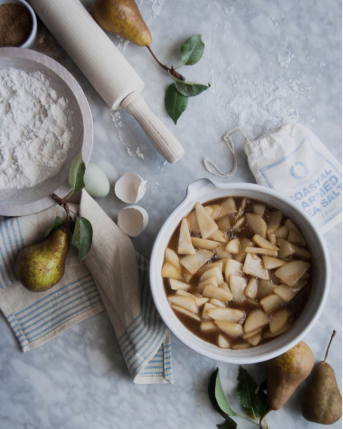 Overhead baking scene with a whitewashed rolling pin, bowl of sliced pears, flour, fresh pears, and sprigs of greenery.