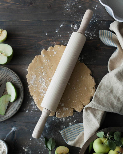 Whitewashed ash rolling pin resting across flour-dusted pastry dough on a dark wooden table surrounded by green apples and baking linens.