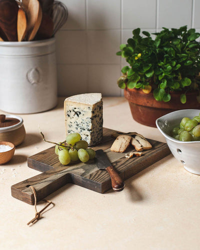Dark wood Pantry Cutting Board styled with cheese, crackers, grapes, and a metal cheese knife beside a potted herb and ceramic bowl.