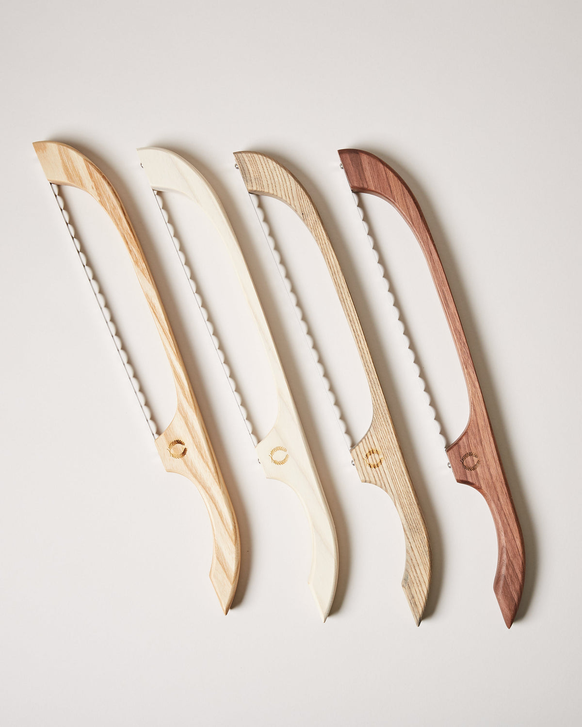 Four curved bread bow knives with serrated metal blades and wooden frames in pale maple, cream, natural oak, and dark walnut finishes.