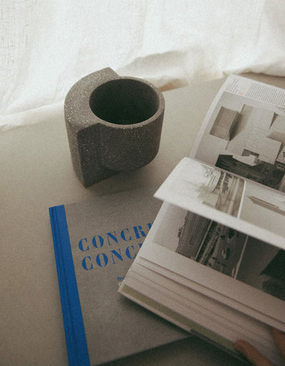Graphite Tortuga Forma Platform Planter Small - Terrazzo beside an open architecture book and stacked books on a light surface against draped white fabric, with its empty circular opening visible from above.
