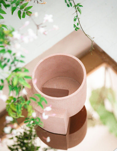 Coral planter from the Tortuga Forma Platform Planter Set viewed from above on a sunlit wooden surface among leafy branches, showing its empty circular interior, speckled finish and projecting slab support.