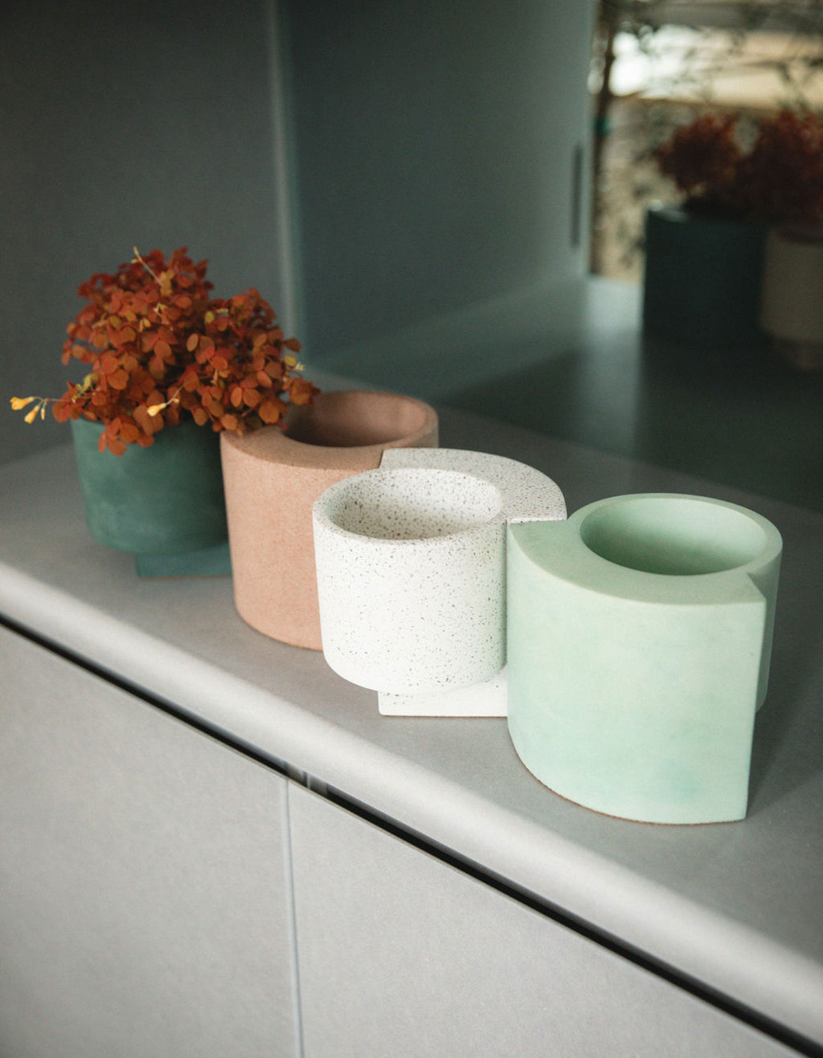 Tortuga Forma Platform Planter Small - Terrazzo displayed among coral, white and pale green planters on a grey cabinet, with orange flowers in a darker planter at the back and sunlight illuminating the empty pots in front.