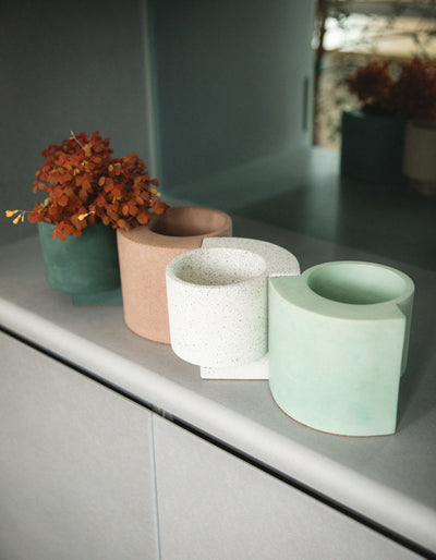 Tortuga Forma Platform Planter Small - Terrazzo displayed among coral, white and pale green planters on a grey cabinet, with orange flowers in a darker planter at the back and sunlight illuminating the empty pots in front.