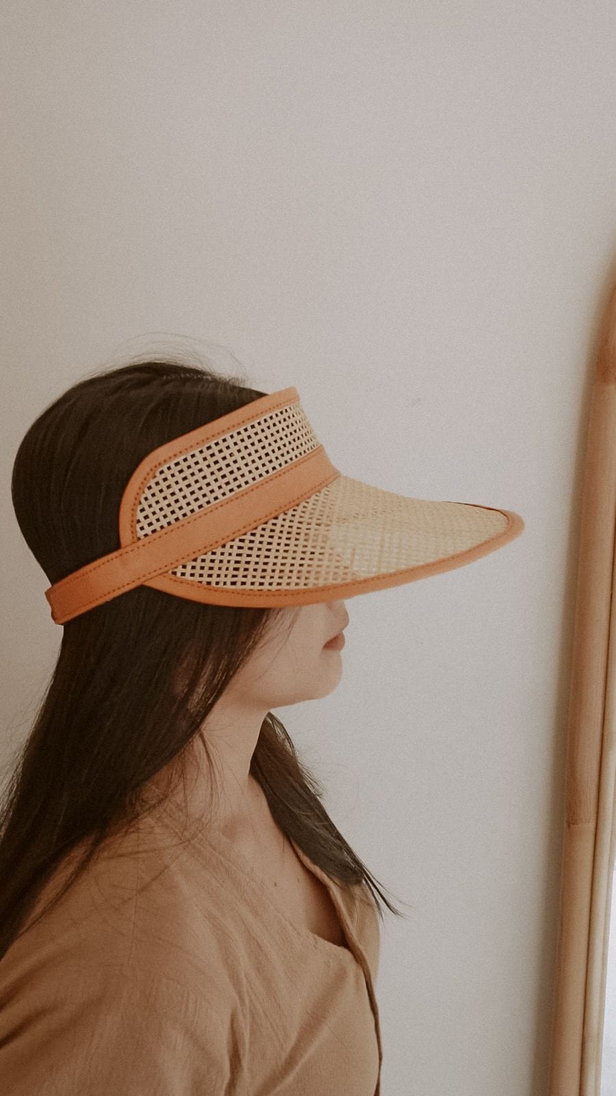 Village Thrive Sumi Leather and Cane Rattan Visor, camel rattan cane with dark leather trim, worn in profile against a wall.