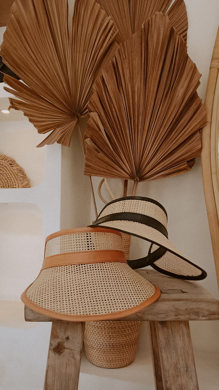 Village Thrive Sumi Leather and Cane Rattan Visor, camel and black rattan cane, two visors displayed on a stand with dried palm fronds.