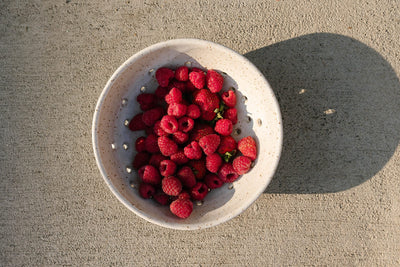 White ceramic colander filled with raspberries on a concrete surface