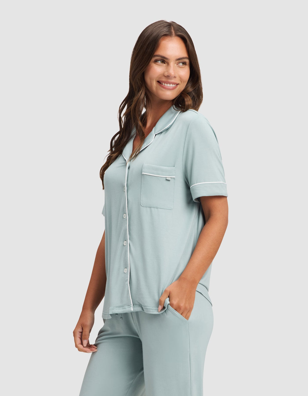 Closer view of the pale blue pajama set, showing the top’s notched collar, buttons, chest pocket, and white piping, with the model’s hand in a pants pocket.