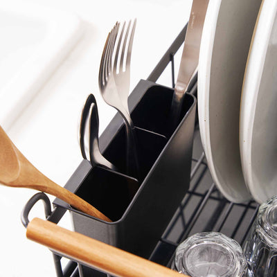 Close-up of the Yamazaki Home Dish Rack - Steel + Wood utensil caddy, showing separate black compartments holding a wooden spoon, cutlery and dark utensils beside upright plates and a wooden handle.