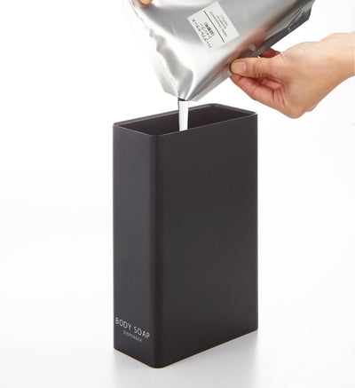 Liquid streams from a silver refill pouch into the black Yamazaki Home Rectangle Shower Dispenser - Three Styles labelled BODY SOAP.