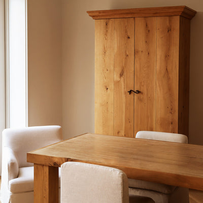 A tall wooden cabinet with two doors and dark metal handles stands in a dining room corner next to a wooden table and cream-colored upholstered chairs by House of Leon.