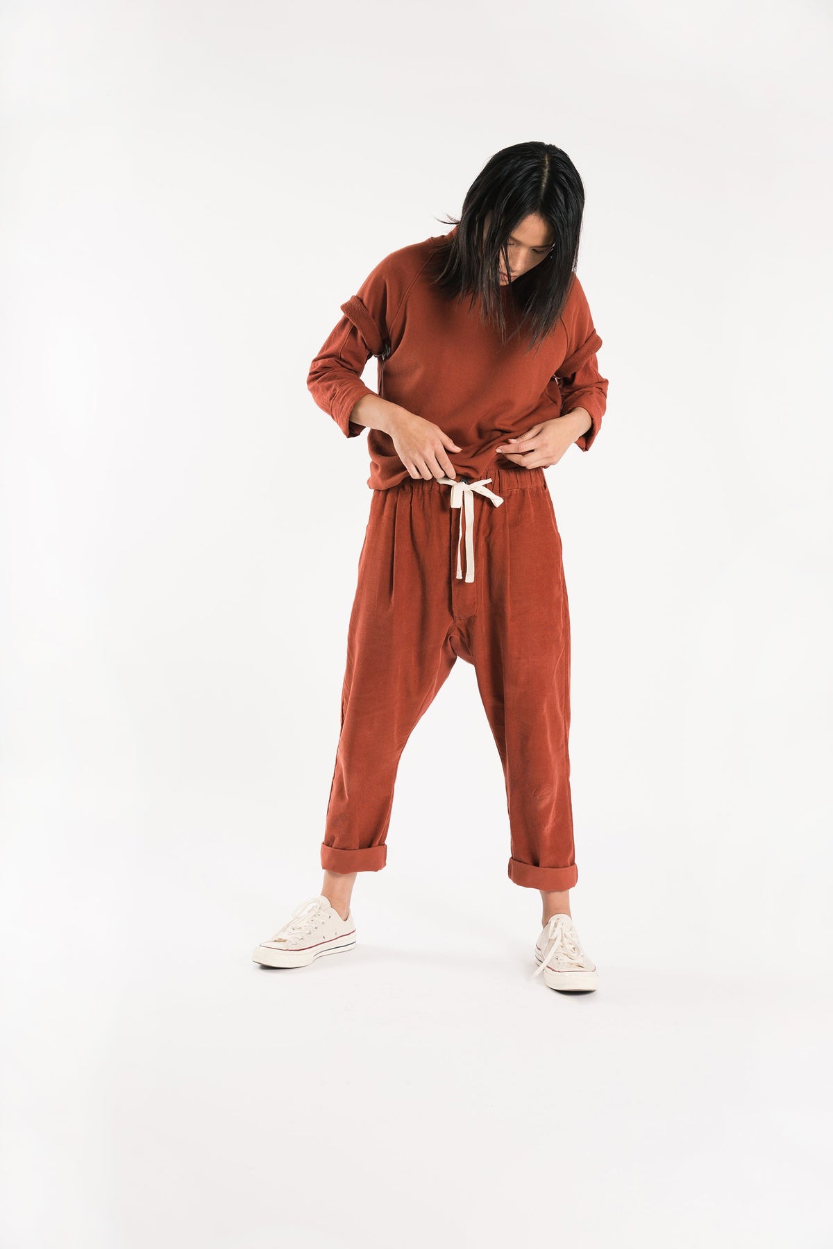 Model adjusting the drawstring of rust-red Paddington pants worn with a matching shirt and white sneakers.