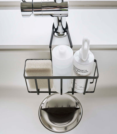 Overhead view of the black Yamazaki Home Faucet-Hanging Sponge Caddy - Steel holding a beige sponge, white dispenser and spray bottle directly above a round sink drain.