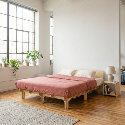 Studio Furnis DOVID natural wood bed frame with dusty rose bedding, a low curved headboard and multiple rounded legs in a bright loft bedroom filled with plants