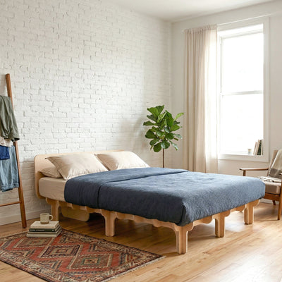 DOVID natural wood bed frame with dark blue bedding in a bright white brick bedroom with a patterned rug and leafy plant