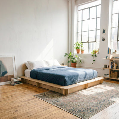 Angled view of a Studio Furnis wood bed platform with blue bedding in a sunlit bedroom with wood floors and a vintage-style rug