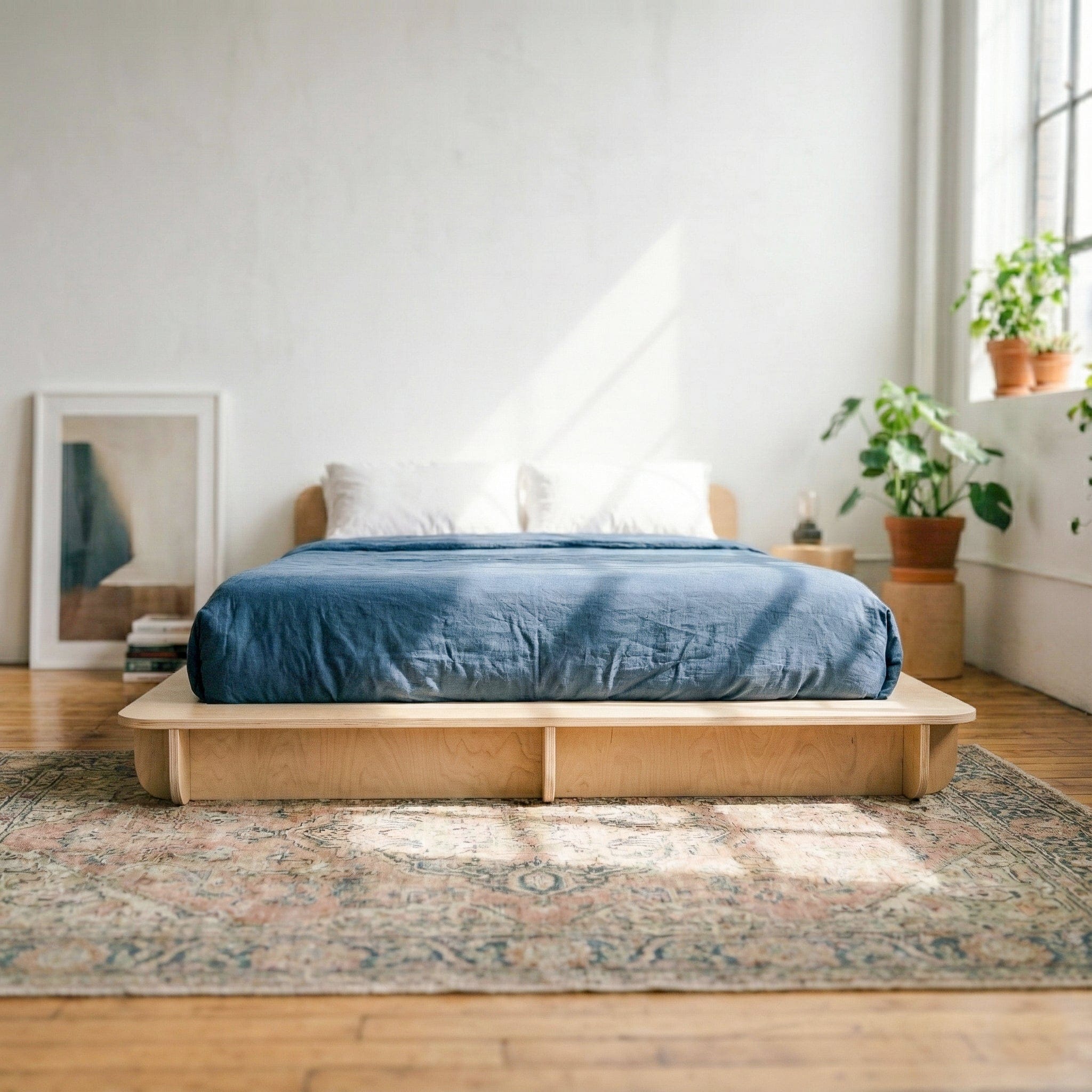 Front view of a Studio Furnis natural wood bed platform with deep blue bedding in a bright white bedroom with plants and a patterned rug