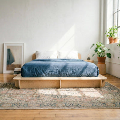 Front view of a Studio Furnis natural wood bed platform with deep blue bedding in a bright white bedroom with plants and a patterned rug
