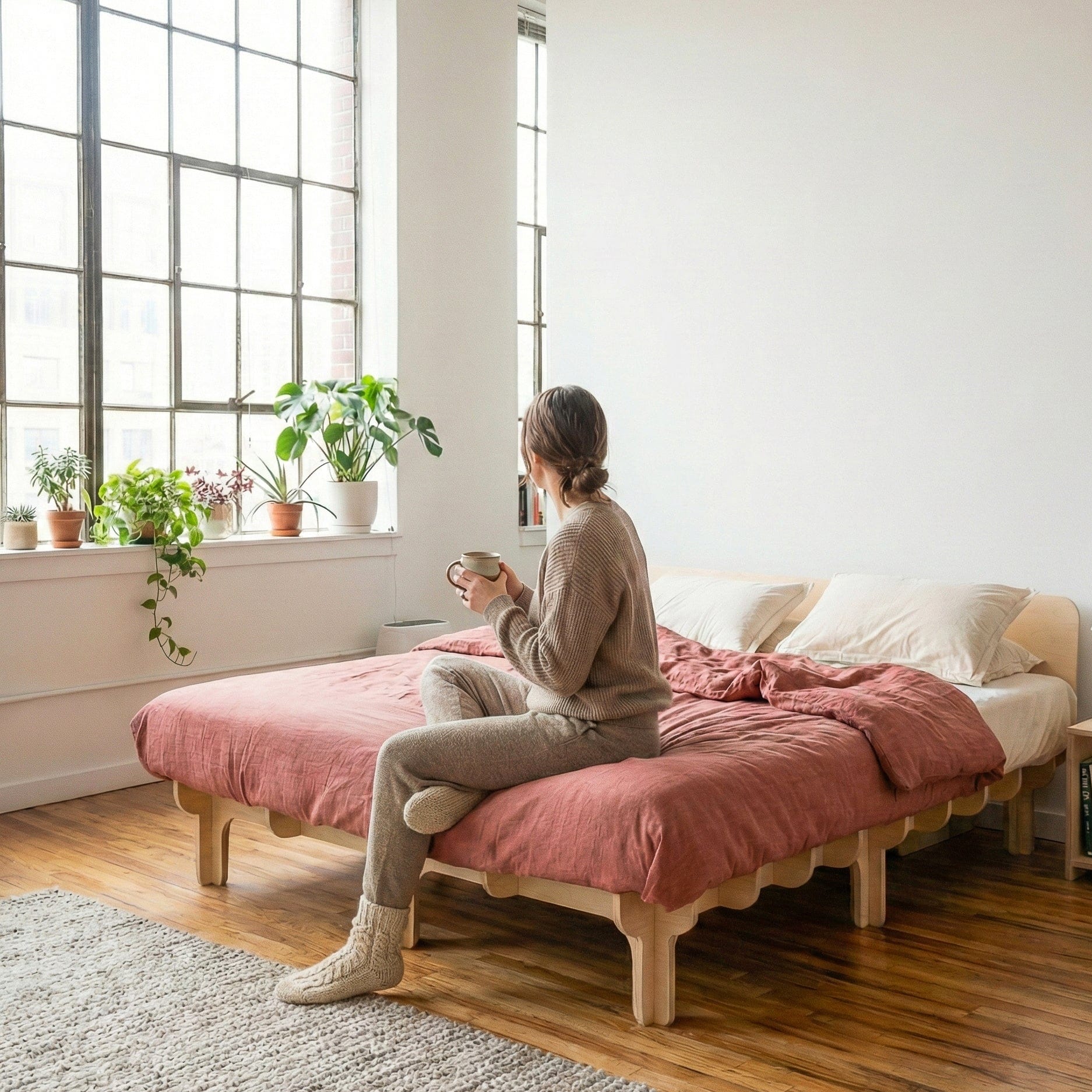 Person seated on the DOVID natural wood bed frame with dusty rose bedding beside large windows and potted plants