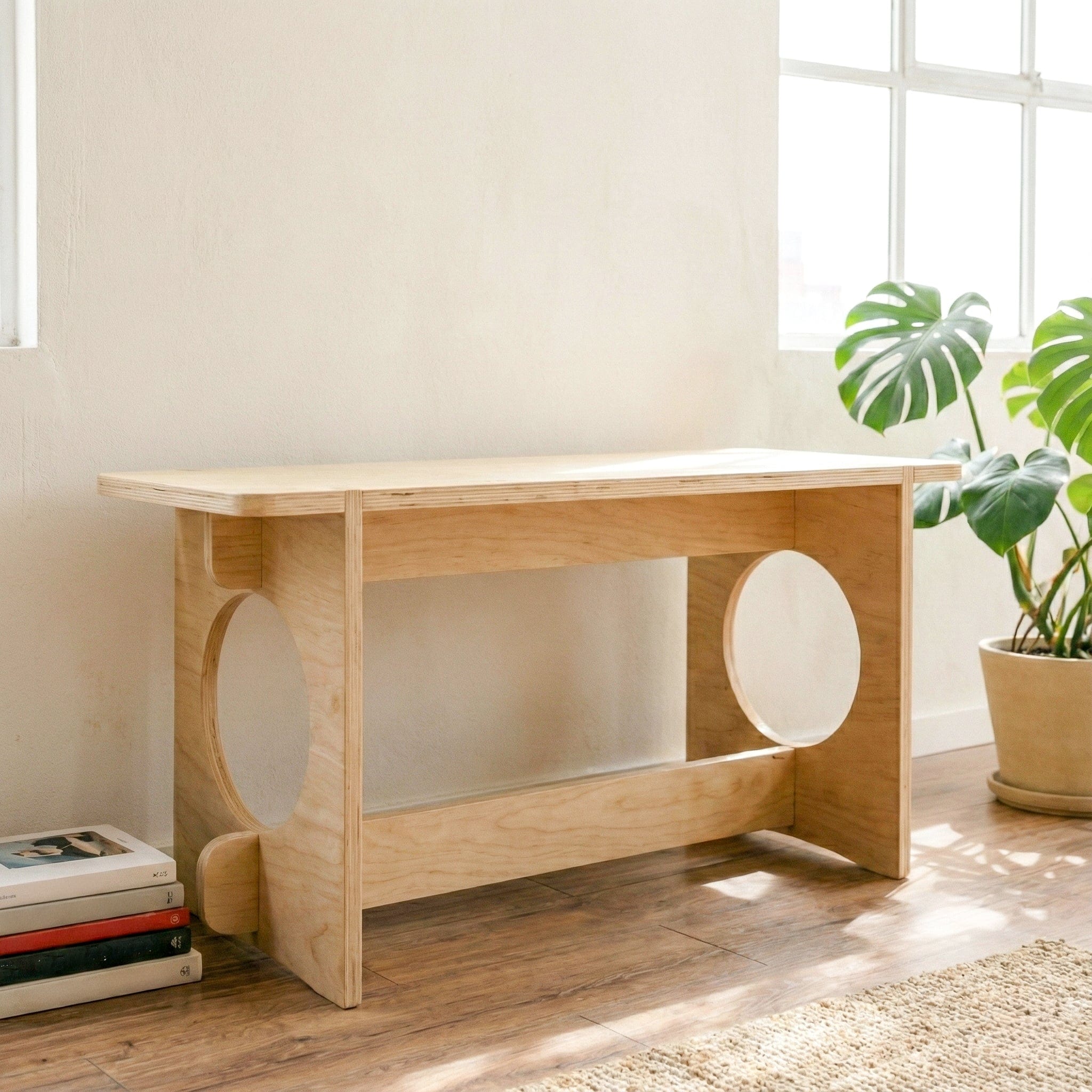 YOYO wood bench in a sunlit room beside a leafy plant and stacked books, showing its pale wood grain and circular side cutouts