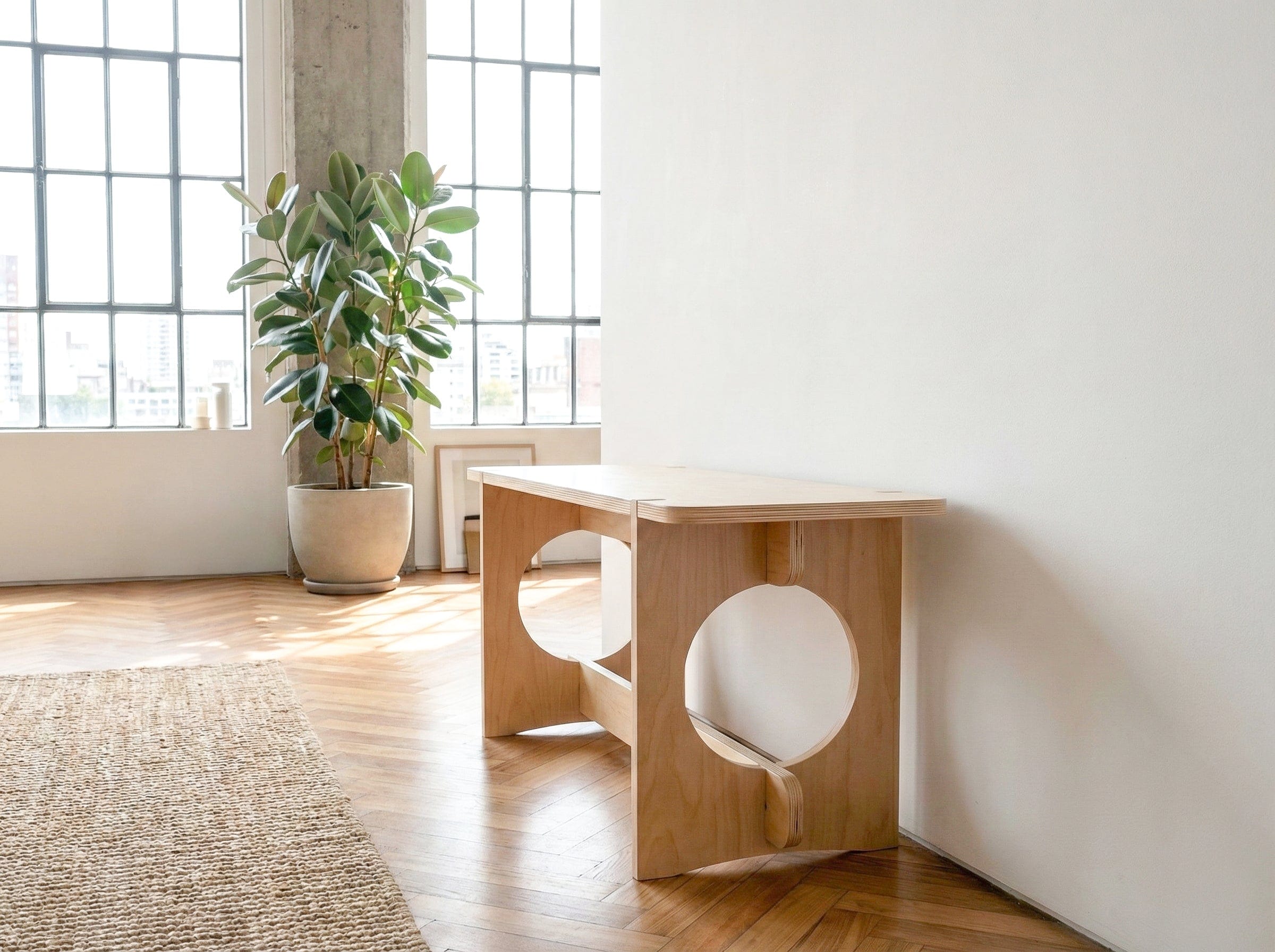 Angled view of the YOYO natural wood bench against a white wall near a potted plant, window and woven rug