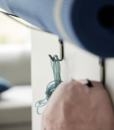 Close-up of the black Yamazaki Home Over-the-Door Fitness Equipment Hanger - Steel hooks beneath a blue mat, with a coiled turquoise jump rope and pink cap hanging from separate hooks.