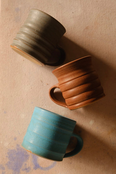 Overhead arrangement of Settle Ceramics The Big Mug in olive-grey, rust brown and turquoise, positioned at different angles on a tan surface to show their ridged sides and rounded handles.