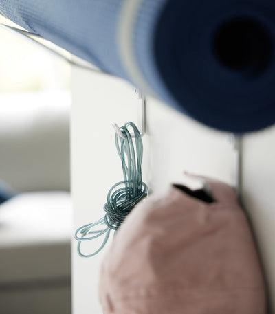 Detail of the white Yamazaki Home Over-the-Door Fitness Equipment Hanger - Steel lower hooks, with a turquoise jump rope and pink cap suspended beneath a rolled blue mat.