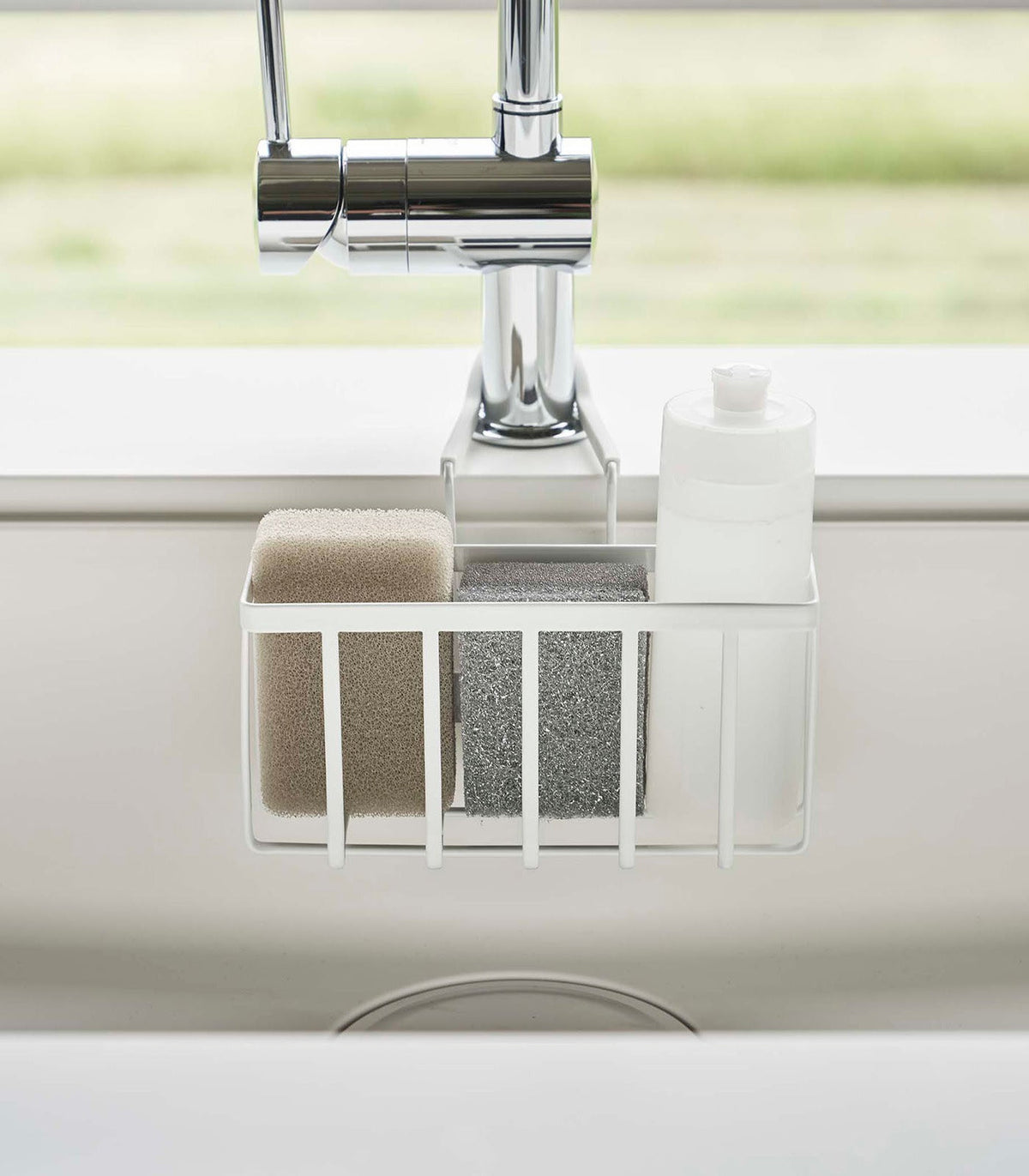 Front view of the white Yamazaki Home Faucet-Hanging Sponge Caddy - Steel holding beige and grey sponges beside a white bottle, with two support arms extending around the faucet.