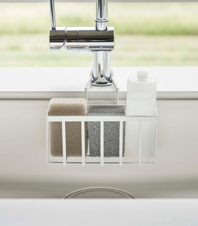 Front view of the white Yamazaki Home Faucet-Hanging Sponge Caddy - Steel holding beige and grey sponges beside a white bottle, with two support arms extending around the faucet.