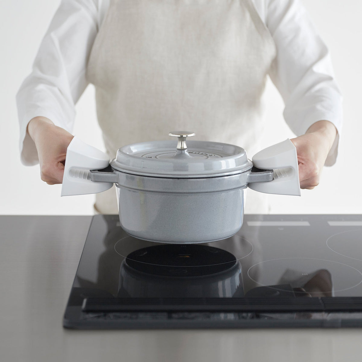 Person wearing a light apron uses white Yamazaki Home Magnetic Pot Holders - Set of 2 - Silicone to grip both handles of a grey lidded pot above a black glass cooktop.
