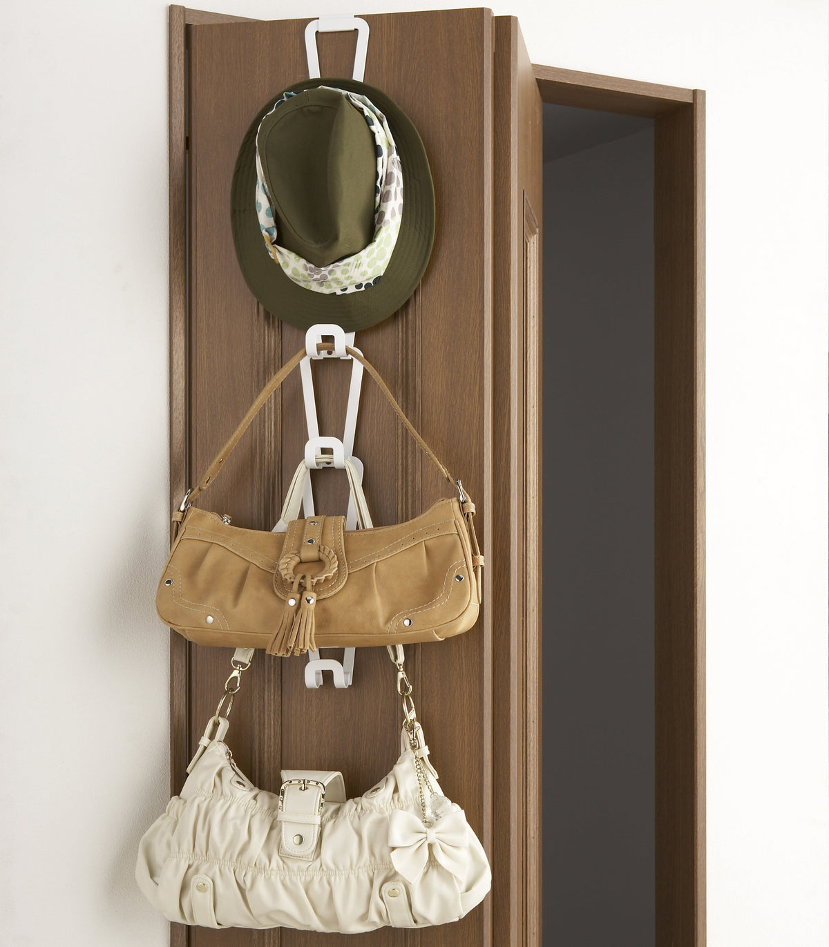 White Yamazaki Home Cascading Storage Hooks - Steel hanging over a wooden door, with linked elongated frames holding an olive hat, tan handbag and cream handbag at different heights.