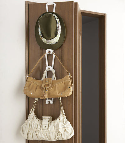 White Yamazaki Home Cascading Storage Hooks - Steel hanging over a wooden door, with linked elongated frames holding an olive hat, tan handbag and cream handbag at different heights.