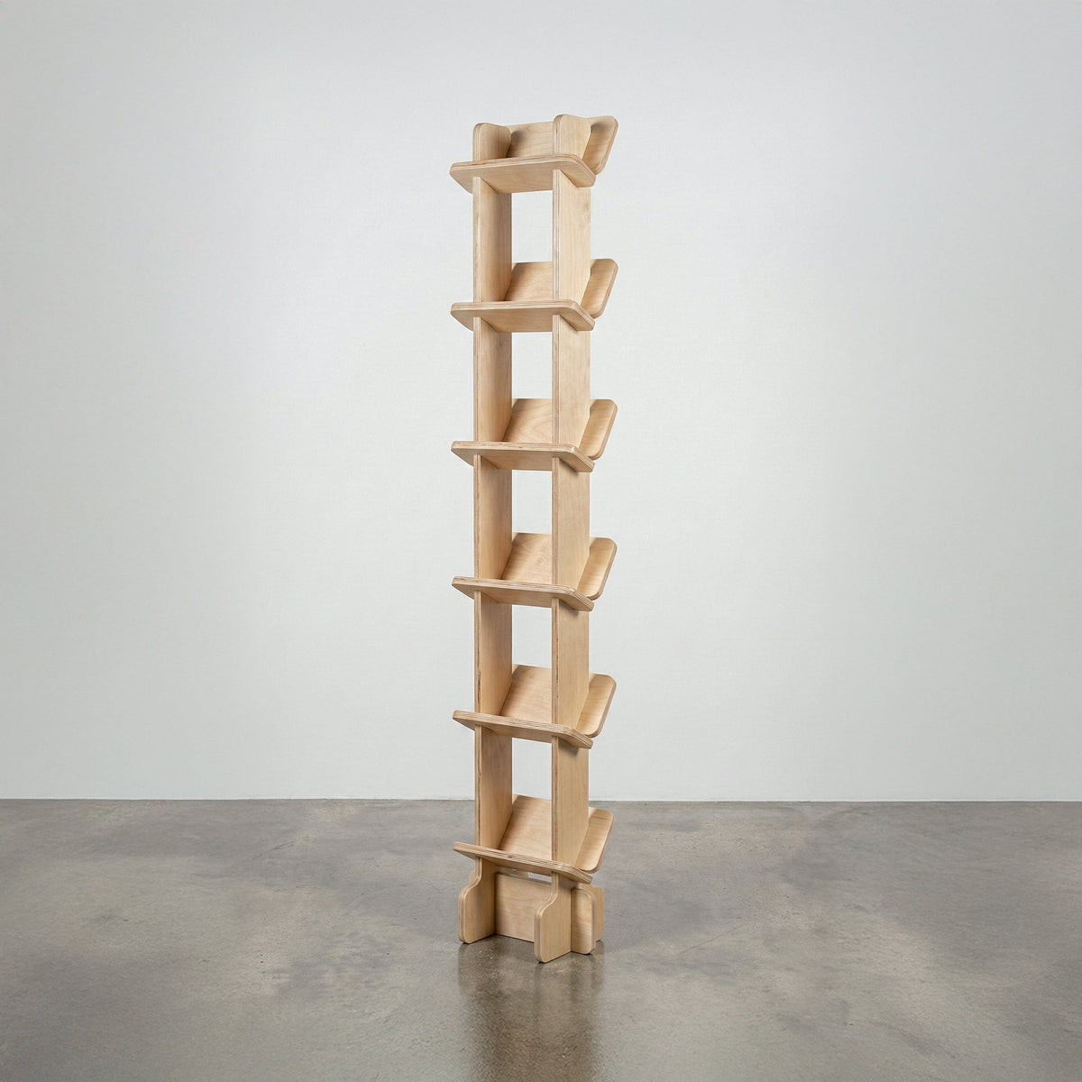 Studio Furnis Yankel tall narrow bookcase in natural wood with a central vertical spine and six pairs of sculptural angled shelves on a concrete floor