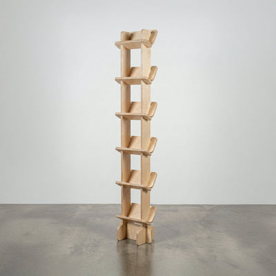 Studio Furnis Yankel tall narrow bookcase in natural wood with a central vertical spine and six pairs of sculptural angled shelves on a concrete floor