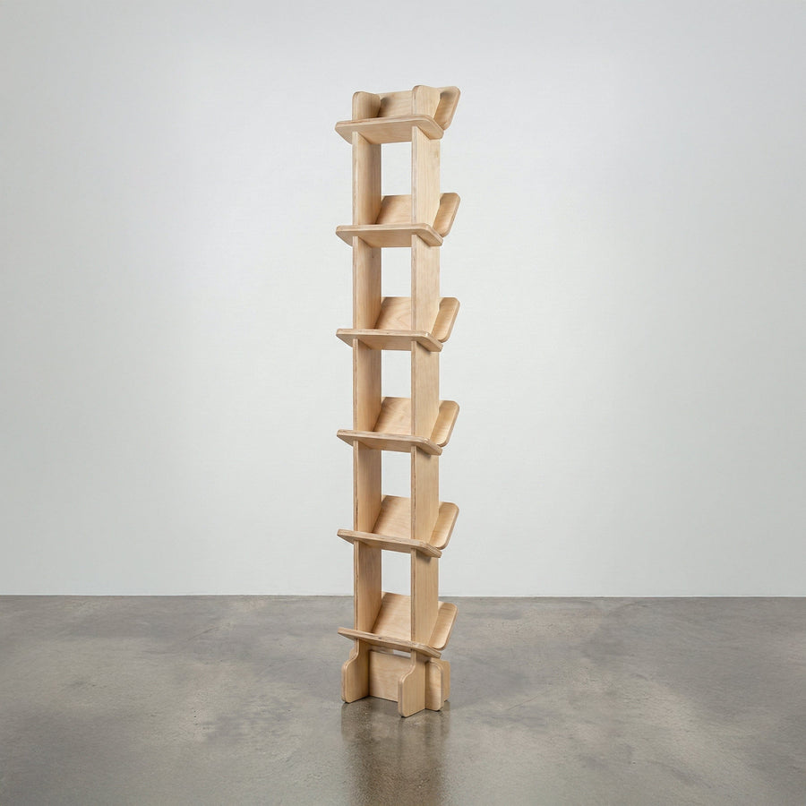 Studio Furnis Yankel tall narrow bookcase in natural wood with a central vertical spine and six pairs of sculptural angled shelves on a concrete floor