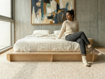 Woman sitting on the edge of a Studio Furnis wood bed platform with white bedding in a minimalist bedroom