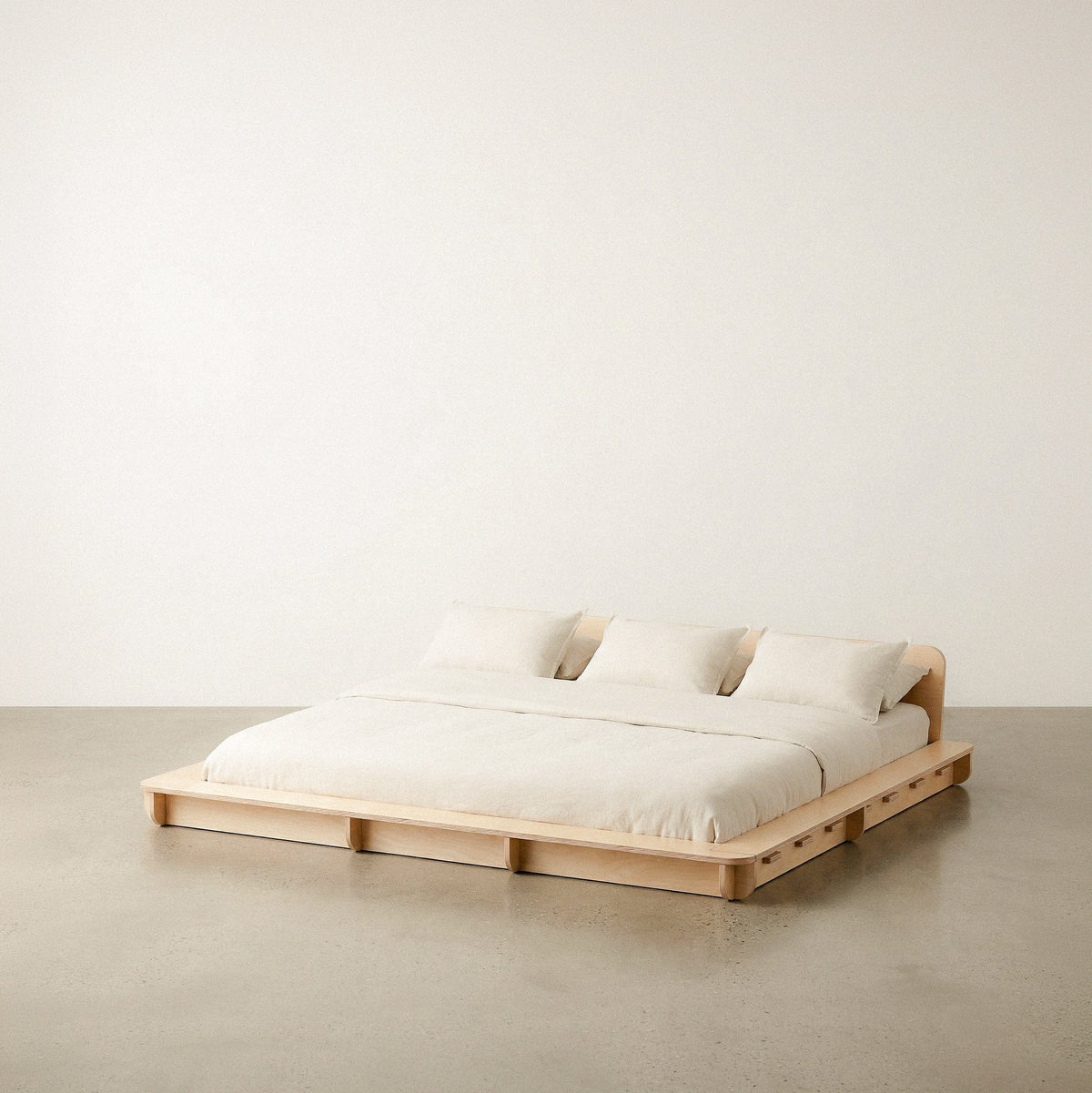 Extra-wide Studio Furnis natural wood bed platform with cream bedding, two pillows and a low curved headboard against a neutral studio background