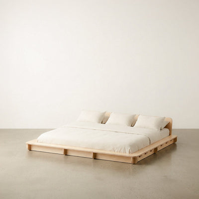 Extra-wide Studio Furnis natural wood bed platform with cream bedding, two pillows and a low curved headboard against a neutral studio background