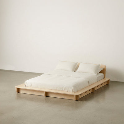 Wide Studio Furnis wood bed platform with cream bedding, one pillow and a low curved headboard against a neutral studio background
