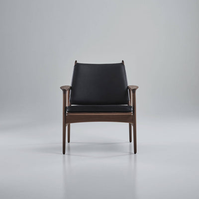 Front view of the black leather Eikund Broadway Lounge Chair with a brown wood frame in a gray studio, showing rounded armrest ends and slender legs.