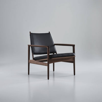 Angled front view of the black leather Eikund Broadway Lounge Chair in a gray studio, showing its open sides, gently reclined backrest, and thin seat cushion above the wood rails.