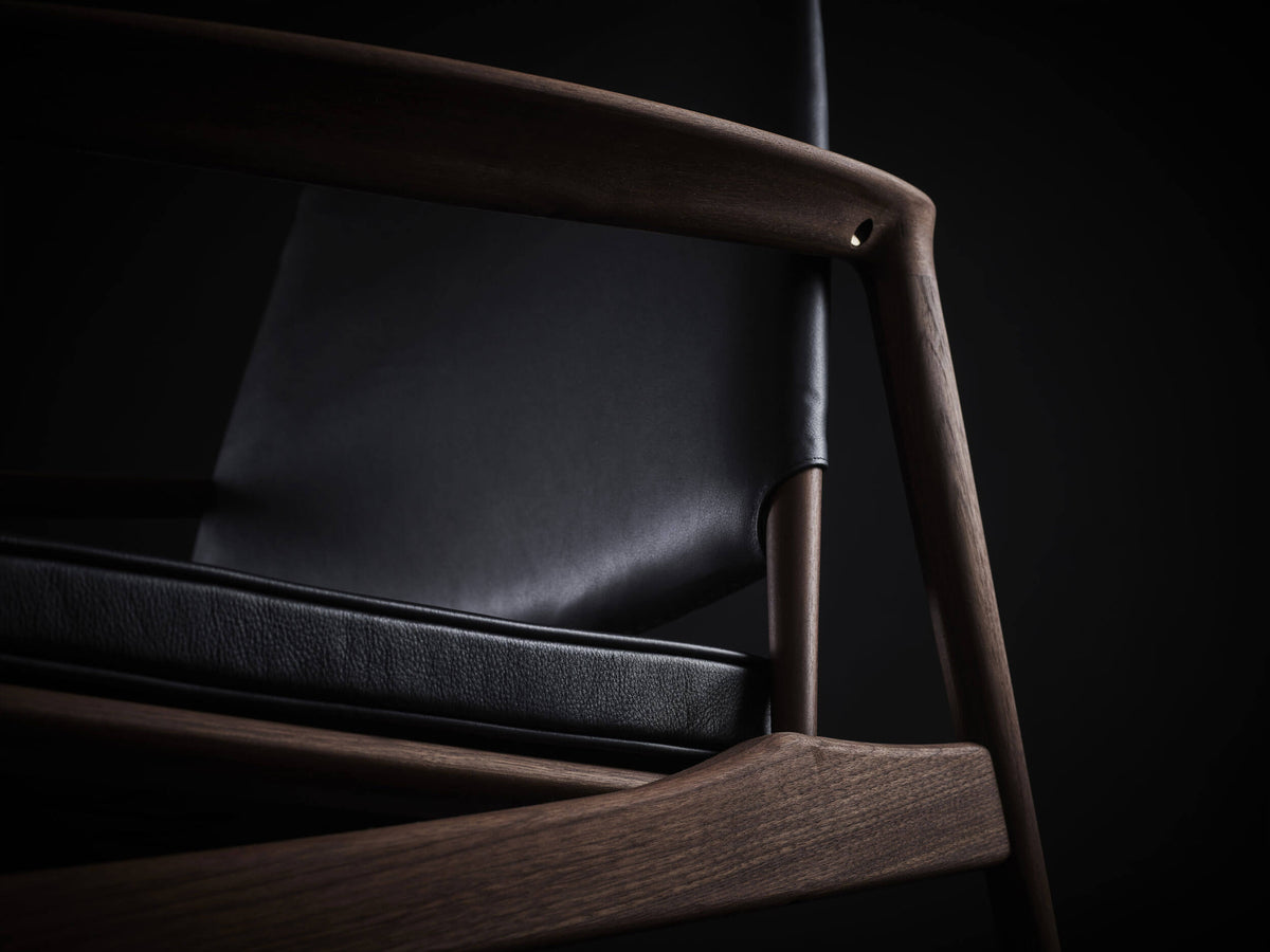 Close-up of the Eikund Broadway Lounge Chair’s dark wood armrest and front support, framing the black leather back and stitched seat cushion.