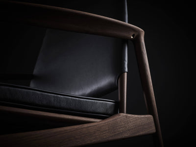 Close-up of the Eikund Broadway Lounge Chair’s dark wood armrest and front support, framing the black leather back and stitched seat cushion.