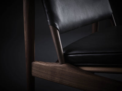 Close-up of the black leather Eikund Broadway Lounge Chair, showing the lower backrest edge, gap above the seat, and grain of the broad wood side rail.