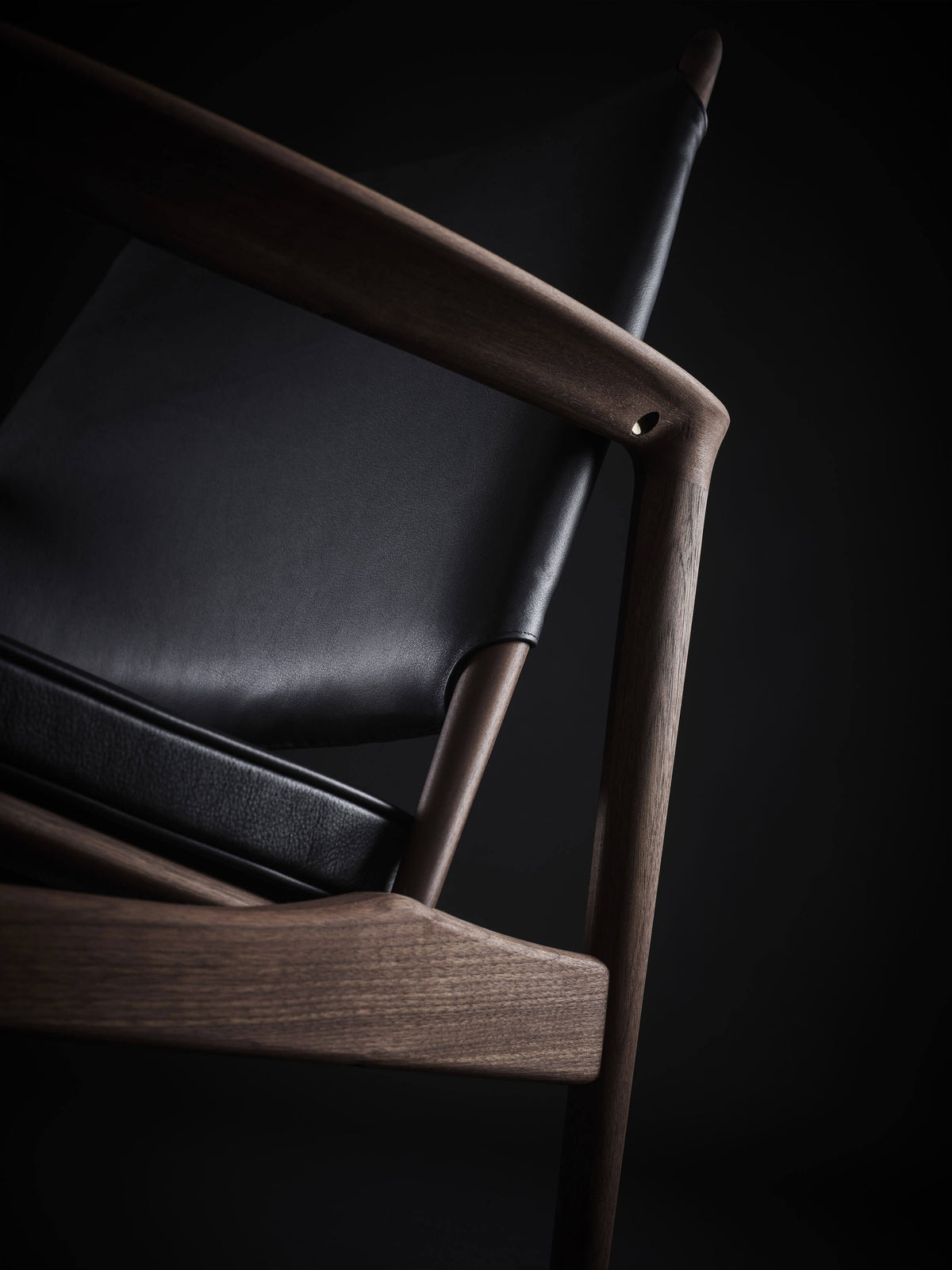 Angled detail of the Eikund Broadway Lounge Chair’s rounded wood armrest joint, black leather seat cushion, and curved wood rail beneath.