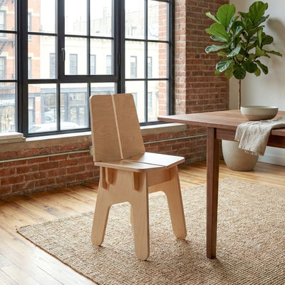 Rear three-quarter view of the BURIJ wood chair beside a dining table in a sunlit loft with exposed brick, plants and large windows