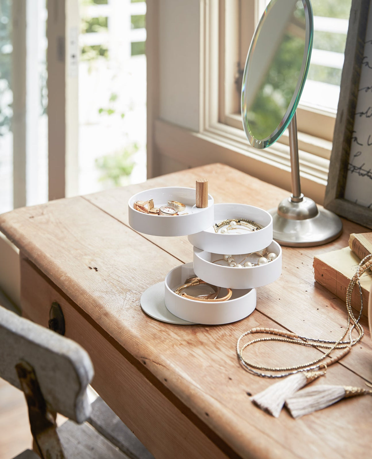 Yamazaki Home Open Top Stacked Jewelry Organizer with its white trays fanned open on a wooden dressing table beside a window and tabletop mirror, with jewellery separated among the tiers.